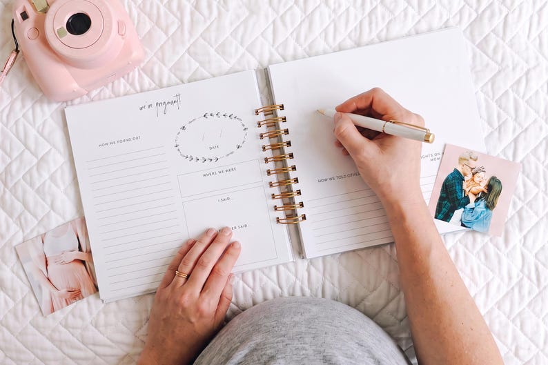 K&ouml;nnte beinhalten: Ein spiralgebundenes Schwangerschaftstagebuch mit wei&szlig;en Seiten, die gedruckte Abschnitte wie "we are pregnant", "how we found out" und "how we told others" enthalten. Eine hellrosa Sofortbildkamera und zwei quadratische Fotos liegen daneben.