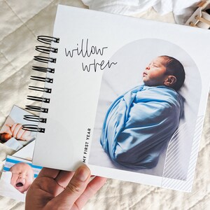 May include: A white baby memory book with a spiral binding. The cover features a photo of a sleeping baby wrapped in a blue blanket and the text "Willow Wren" and "My First Year".