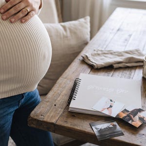 Pregnant woman standing next to table with almond colored "Growing You" pregnancy journal on table surrounded by pregnancy photos and cup of coffee