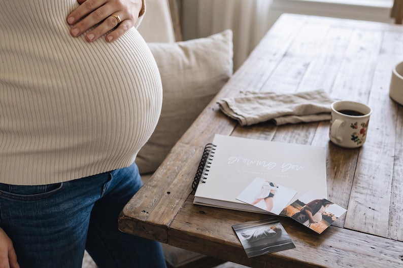 Pregnant woman standing next to table with almond colored "Growing You" pregnancy journal on table surrounded by pregnancy photos and cup of coffee