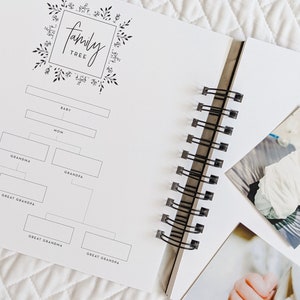 May include: A white family tree journal with a black floral design and the words "family tree" in a black script font. The journal has a spiral binding and is open to a page with a blank family tree chart. The next page has a photo of a baby in a white onesie.
