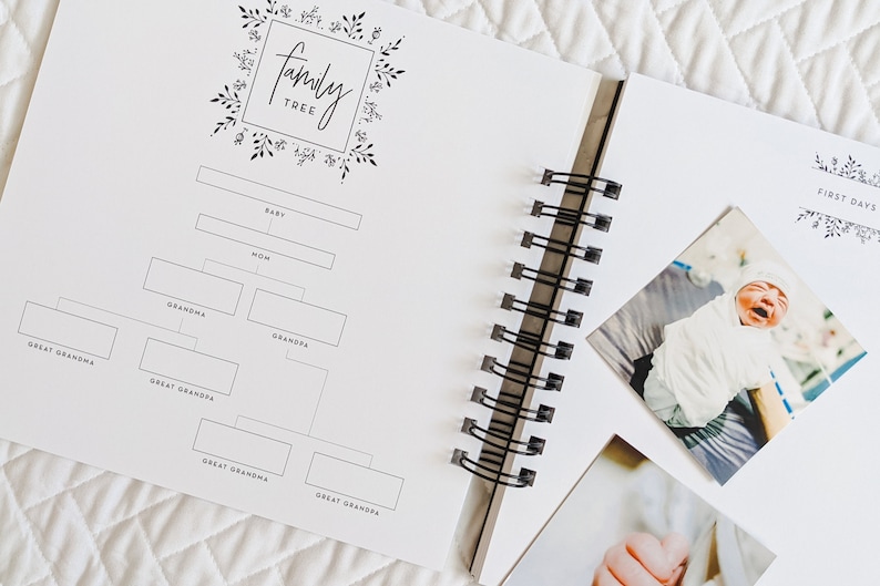 May include: A white family tree journal with a black floral design and the words "family tree" in a black script font. The journal has a spiral binding and is open to a page with a blank family tree chart. The next page has a photo of a baby in a white onesie.
