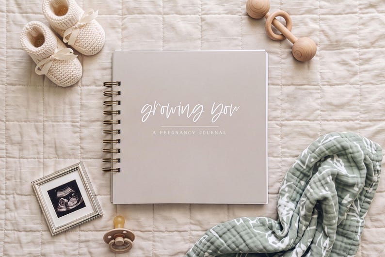 Growing You Pregnancy Journal with almond color cover laying on a bed surrounded by baby items