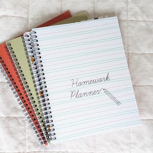 Può includere: Tre quaderni a spirale impilati. Il quaderno superiore ha una copertina bianca con linee orizzontali blu e rosse, il testo "Homework Planner" in corsivo nero e un piccolo disegno al tratto nero di una matita accanto al testo.