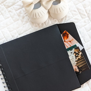 Puede incluir: Un cuaderno negro de espiral con un sobre negro unido a la portada. El sobre tiene un bolsillo para guardar fotos u otros art&iacute;culos. Un bol&iacute;grafo blanco y un par de zapatos de beb&eacute; blancos son visibles en el fondo.