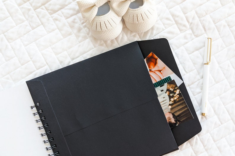 Puede incluir: Un cuaderno negro de espiral con un sobre negro unido a la portada. El sobre tiene un bolsillo para guardar fotos u otros art&iacute;culos. Un bol&iacute;grafo blanco y un par de zapatos de beb&eacute; blancos son visibles en el fondo.