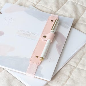 Może przedstawiać: Spiralny dziennik ciążowy zatytułowany Welcoming Baby z okładką w abstrakcyjne akwarelowe wzory w delikatnych odcieniach. Jasnoróżowy elastyczny uchwyt na długopis jest przymocowany do okładki i trzyma biały długopis ze złotymi akcentami.
