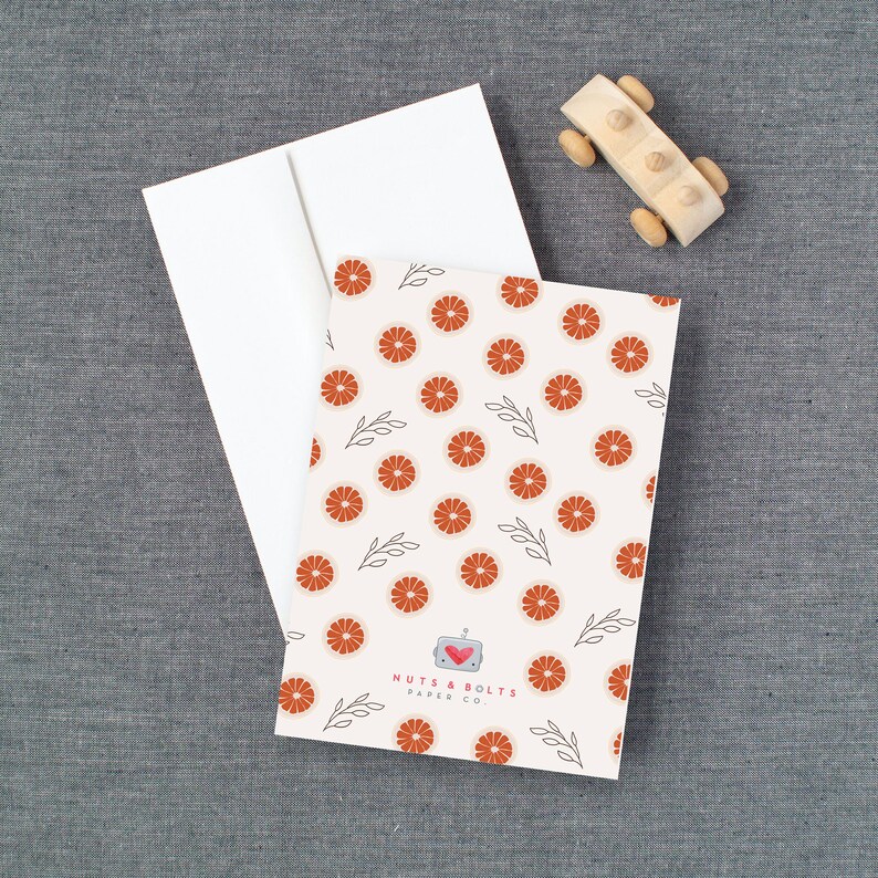 May include: A white card with a red and white grapefruit pattern. The card has the text "Nuts & Bolts Paper Co." in black text.