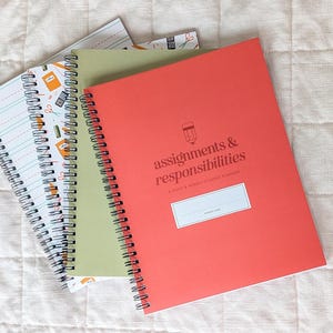 May include: Three spiral-bound notebooks stacked together. The top notebook is coral with the text "assignments & responsibilities" and a pencil icon. The middle notebook is olive green. The bottom notebook features a white cover with school supply patterns.
