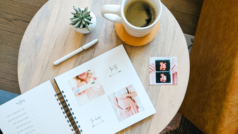 Puede incluir: Un diario de embarazo blanco en espiral con un bol&iacute;grafo negro y una taza de caf&eacute; sobre una mesa de madera. El diario tiene una secci&oacute;n para "The First" e incluye fotos de una ecograf&iacute;a y una mujer embarazada.
