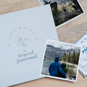 May include: A light blue travel journal with a spiral binding. The cover features a mountain range illustration and the text "Jason & Kelly 2023 our travel journal".