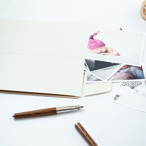 May include: A white spiral bound photo album with a pocket on the front cover. The album is open and there are several photographs inside, including a photo of a baby wearing a pink hat. There is also a wooden bead necklace and a pen on the table.