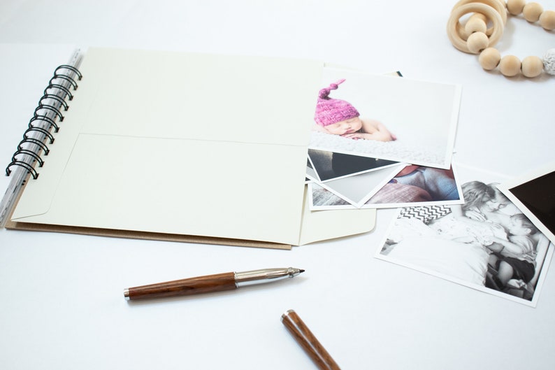 May include: A white spiral bound photo album with a pocket on the front cover. The album is open and there are several photographs inside, including a photo of a baby wearing a pink hat. There is also a wooden bead necklace and a pen on the table.