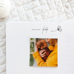 Puede incluir: Un &aacute;lbum de fotos blanco con una encuadernaci&oacute;n en espiral negra. La portada tiene el texto "Your First Photo" y una simple ilustraci&oacute;n de diente de le&oacute;n. Una foto de una madre sosteniendo a un beb&eacute; reci&eacute;n nacido est&aacute; insertada en el &aacute;lbum.