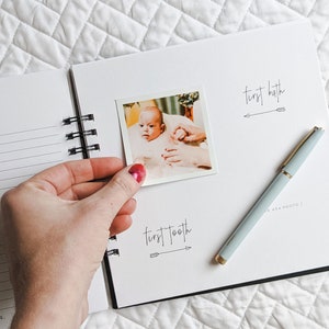 May include: A white baby memory book with a black spiral binding. The book has a photo of a baby in a bath tub and the text "first bath" and "first tooth" with arrows.
