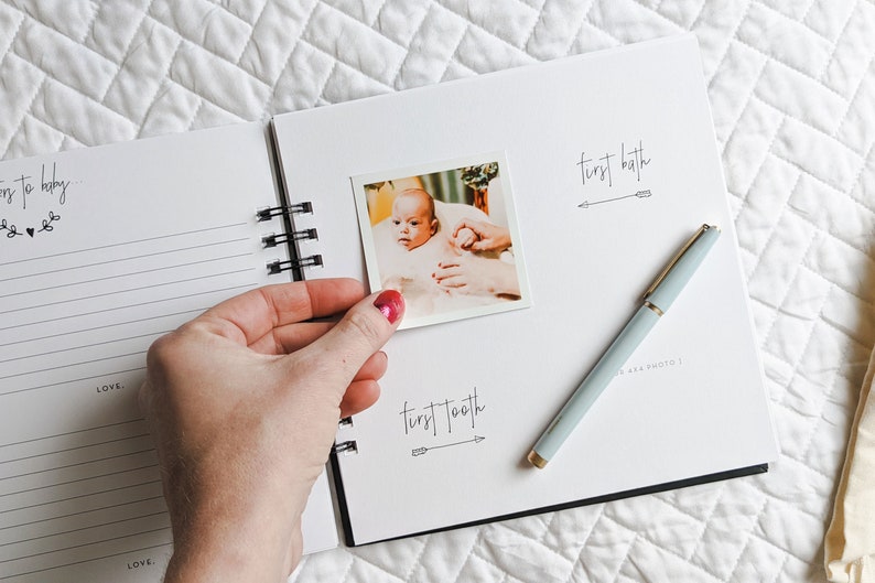 May include: A white baby memory book with a black spiral binding. The book has a photo of a baby in a bath tub and the text "first bath" and "first tooth" with arrows.