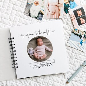 May include: A white baby photo album with a spiral binding. The cover features a circular cutout with a photo of a baby in a pink outfit. The text "welcome to the world" is written in black cursive script above the photo. The baby's name "JACK OWEN" is written in black text below the photo.