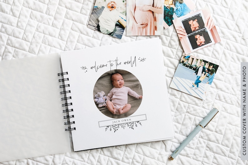 May include: A white baby photo album with a spiral binding. The cover features a circular cutout with a photo of a baby in a pink outfit. The text "welcome to the world" is written in black cursive script above the photo. The baby's name "JACK OWEN" is written in black text below the photo.