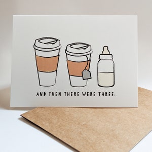 May include: A white card with a black and white illustration of two coffee cups and a baby bottle. The text "AND THEN THERE WERE THREE." is printed below the illustration.