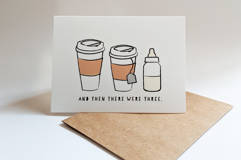 May include: A white card with a black and white illustration of two coffee cups and a baby bottle. The text "AND THEN THERE WERE THREE." is printed below the illustration.