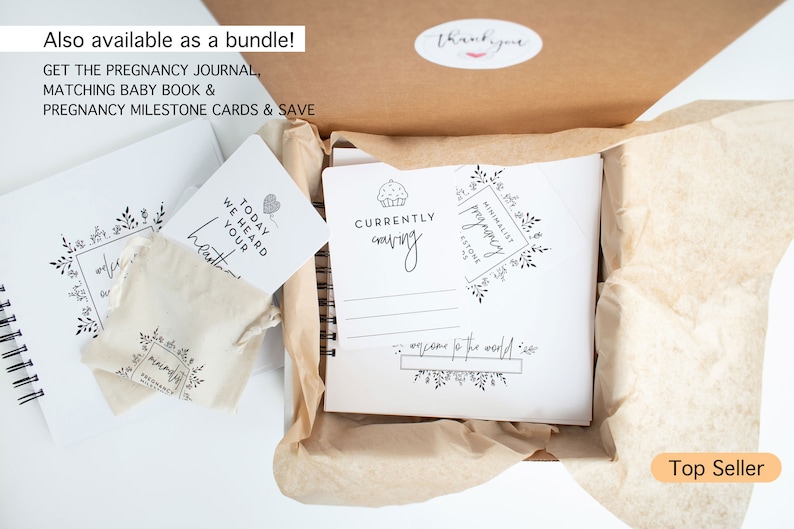May include: A white box with a brown paper liner, containing a pregnancy journal, a baby book, and a set of milestone cards. The journal has the text "Currently craving" and "Minimalist Pregnancy Journal" on the cover. The box is labeled "Top Seller".