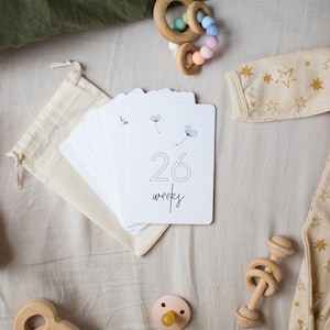 "26 Weeks" Minimalist Pregnancy Milestone Card laying on bed surrounded by baby items by Nuts & Bolts Paper Co