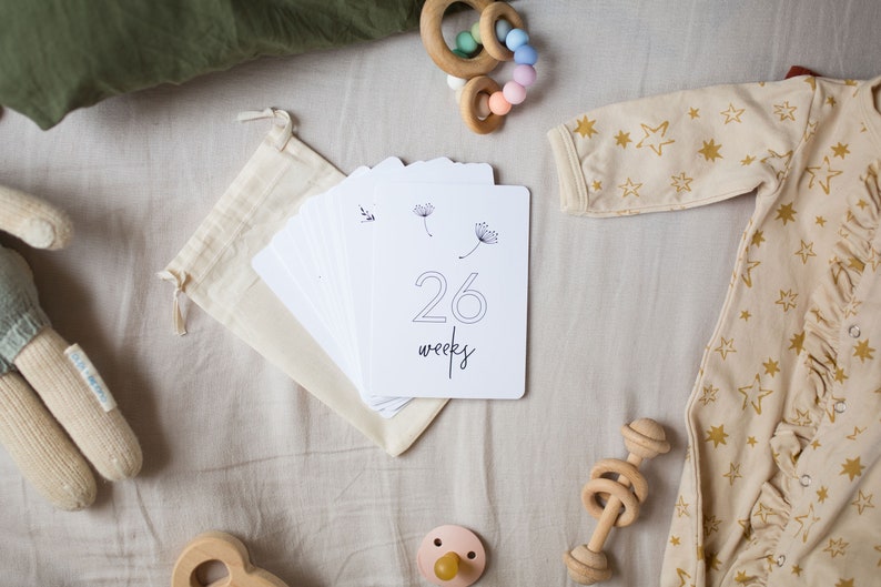 "26 Weeks" Minimalist Pregnancy Milestone Card laying on bed surrounded by baby items by Nuts & Bolts Paper Co