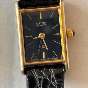 May include: A rectangular Citizen Quartz watch with a gold-tone case and a black dial. The face features gold-tone hour markers and hands, with the text CITIZEN QUARTZ printed in gold. It includes a black textured leather strap and a gold crown.
