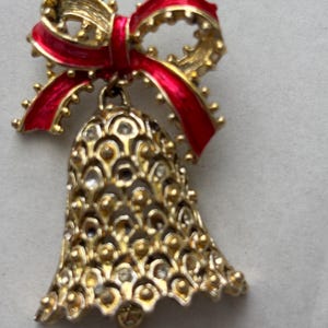 May include: A gold-tone metal brooch shaped like a bell with a textured, openwork pattern and small clear rhinestones. The bell is topped with a bright red enameled bow that features a textured gold-tone border and a small dangling clapper.