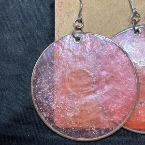 May include: A pair of circular earrings featuring translucent, textured pink material encased in a thin metallic rim. The earrings hang from silver-toned metal hooks attached to a brown cardboard display card with a floral pattern at the top edge.