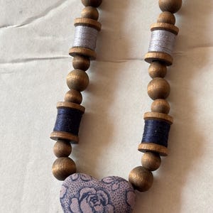 Puede incluir: Un collar con cuentas de madera redondas de color marrón intercaladas con carretes de madera envueltos en hilo gris claro y azul oscuro. La pieza central es un colgante en forma de corazón cubierto de tela morada clara con rosas azul oscuro.