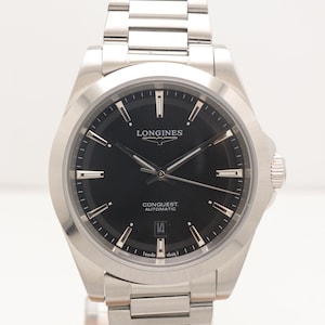 May include: A silver-tone stainless steel Longines Conquest Automatic watch with a black dial, silver hour markers, and a date window at the six o'clock position. The watch features a matching link bracelet and the text Longines Conquest Automatic.