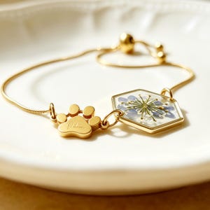 May include: A gold-toned adjustable bracelet featuring a small metal paw print charm with an engraved name and a hexagonal pendant containing a pressed blue flower encased in clear resin, connected by a delicate gold-toned snake chain.
