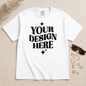 Può includere: Una t-shirt bianca a maniche corte con il testo nero "YOUR DESIGN HERE" stampato in un carattere ondulato sul petto, accentuato da due icone a stella a quattro punte sopra e sotto il testo. La maglietta ha un girocollo e un'etichetta Gildan.