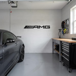 Pode incluir: Uma placa preta montada na parede com o logótipo AMG, apresentando as suas barras inclinadas características e letras em negrito. A placa está exposta numa parede de blocos de betão branco dentro de uma garagem, sobre uma bancada de madeira.