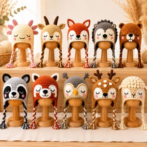 May include: A collection of ten handmade crochet animal hats displayed on wooden stands. The hats feature various designs including an axolotl, deer, fox, hedgehog, bear, raccoon, red panda, owl, reindeer, and sheep, all with braided earflaps.