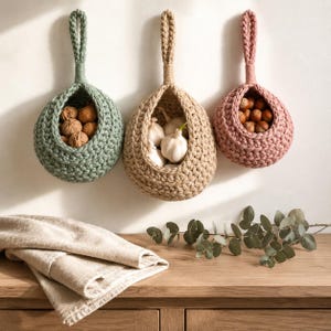 May include: Three teardrop-shaped hanging baskets made of thick, chunky knit cord in sage green, beige, and dusty rose. Each basket features a sturdy loop for wall mounting and contains food items: walnuts, garlic bulbs, and hazelnuts respectively.