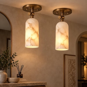 May include: Two ceiling-mounted light fixtures featuring cylindrical shades with a white and tan marbled pattern. Each lamp has a circular brass-toned base and a swivel joint connector, emitting a warm glow from the open bottom end.