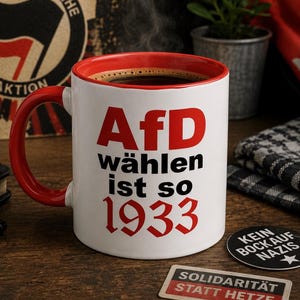 Könnte beinhalten: Eine weiße Keramiktasse mit rotem Innenraum und rotem Henkel, gefüllt mit schwarzem Kaffee. Auf der Vorderseite ist der Text 'AfD wählen ist so 1933' in roter und schwarzer Fettschrift aufgedruckt. Die Tasse steht auf einer dunklen Holzoberfläche.