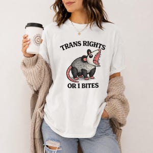 Könnte beinhalten: Ein weißes T-Shirt mit kurzen Ärmeln und einer Grafik eines grau-weißen Opossums mit geöffnetem Maul. Der Text über dem Opossum lautet TRANS RIGHTS und der Text darunter OR I BITES in fetter schwarzer Schrift. Das Shirt hat einen Rundhalsausschnitt.