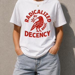Könnte beinhalten: Ein weißes kurzärmeliges T-Shirt mit einem roten Grafikdruck eines Vogels, der einen Zweig hält. Der Text "RADICALIZED BY BASIC DECENCY" ist in Rot gedruckt und wölbt sich über und unter der Vogelillustration. Kombiniert mit einer hellblauen Jeans.