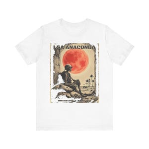 May include: A white t-shirt featuring a graphic print of a skeleton in a military helmet sitting on sandbags. The scene includes a large red moon, palm trees, and the text LSA ANACONDA and MORTARITAVILLE: BALAD, IRAQ in a distressed style.