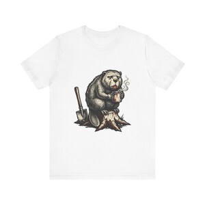 May include: A white t-shirt featuring a graphic illustration of a beaver sitting on a tree stump. The beaver is holding a steaming mug, and a shovel is positioned in the ground next to the stump. The design uses shades of brown, grey, and black.