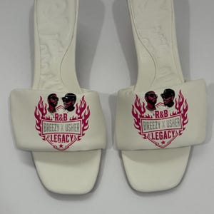 May include: A pair of white open-toe slide sandals with a square toe and a low heel. The wide strap features a graphic print of two men, pink flames, and the text R&B BREEZY X USHER LEGACY. The footbed has a molded design with a subtle embossed pattern.