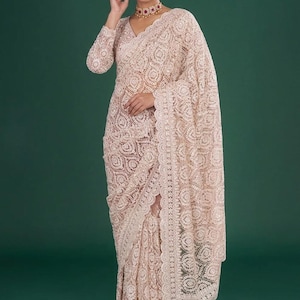 May include: A pale peach-colored saree featuring intricate white floral embroidery throughout the fabric. The garment includes a long-sleeved blouse with a matching pattern, a scalloped border edge, and a sheer, lightweight texture throughout the design.