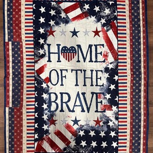 May include: A white, blue, and red patriotic quilt with a distressed American flag design. The quilt features the words "Home of the Brave" in a blue font with a red heart in the center. The quilt is surrounded by a border of red, white, and blue stripes and stars.