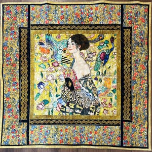 May include: A square quilt featuring a central image of a woman in a decorative dress, surrounded by floral and peacock imagery. The quilt has a yellow, black, and gold border with a floral pattern. The overall design is reminiscent of a painting.
