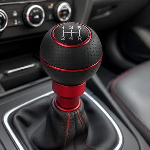 May include: A spherical manual gear shift knob featuring a black perforated leather texture with a red metallic ring accent. The top displays a white gear pattern with numbers 1, 3, 5, 2, 4, and R. It is mounted on a red cylindrical base and black boot.