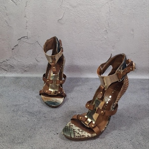 Könnte beinhalten: Ein Paar hochhackige Sandalen mit einer braun-grünen Schlangenledermuster-Textur, braunen Lederriemen mit goldfarbenen Metallakzenten und einer metallisch goldenen Knöchelmanschette mit Schnallenverschluss. Das Design umfasst eine T-Riemen-Konstruktion.