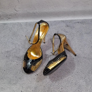 May include: A pair of high-heeled shoes featuring a black patent leather exterior with metallic gold accents on the toe and heel. The shoes include a thin ankle strap with a buckle closure and a gold-toned interior lining with a stiletto heel.